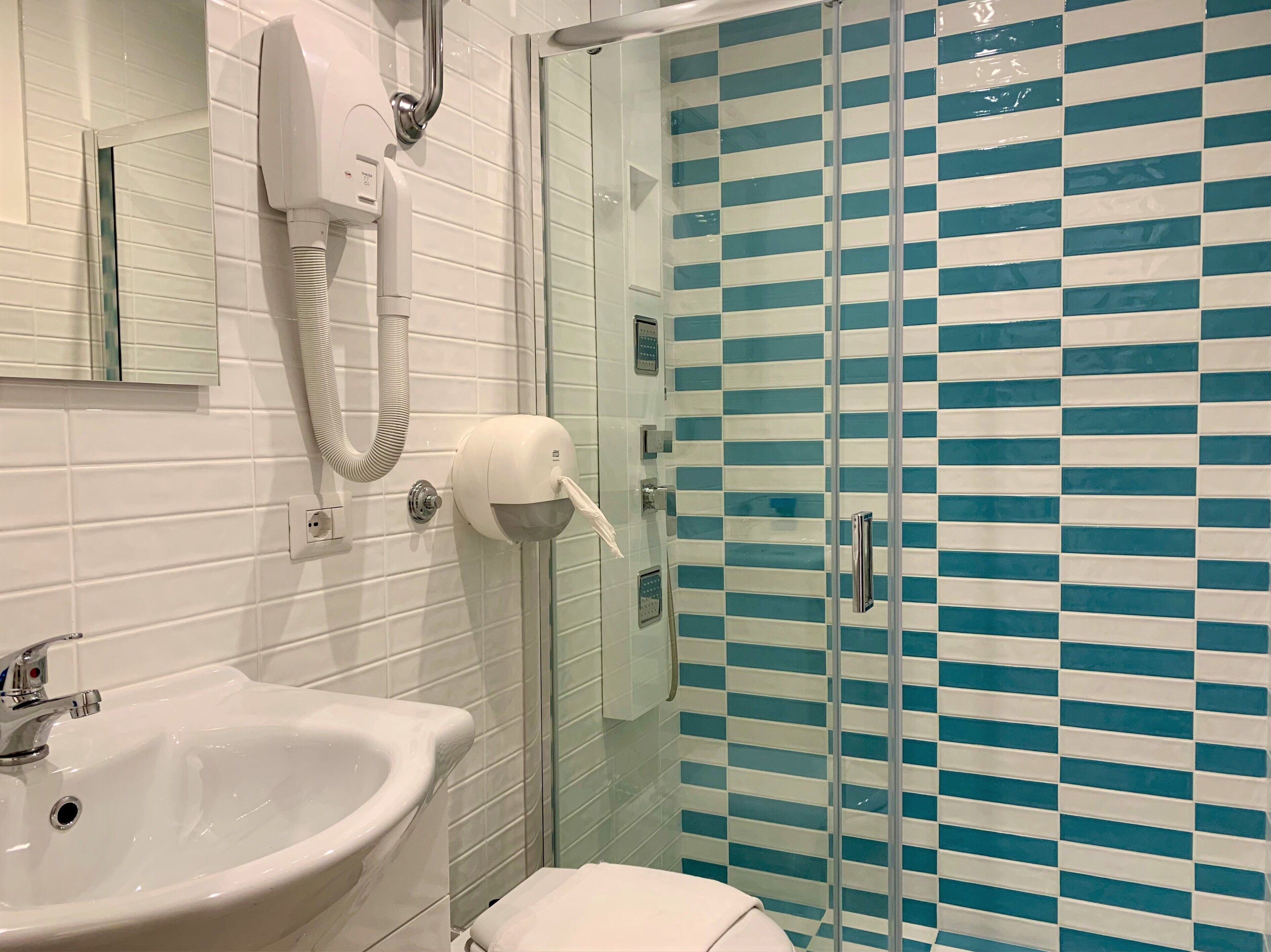 A bathroom with a white sink, a towel rack, a hand dryer, and a glass shower enclosure with blue and white tiles.
