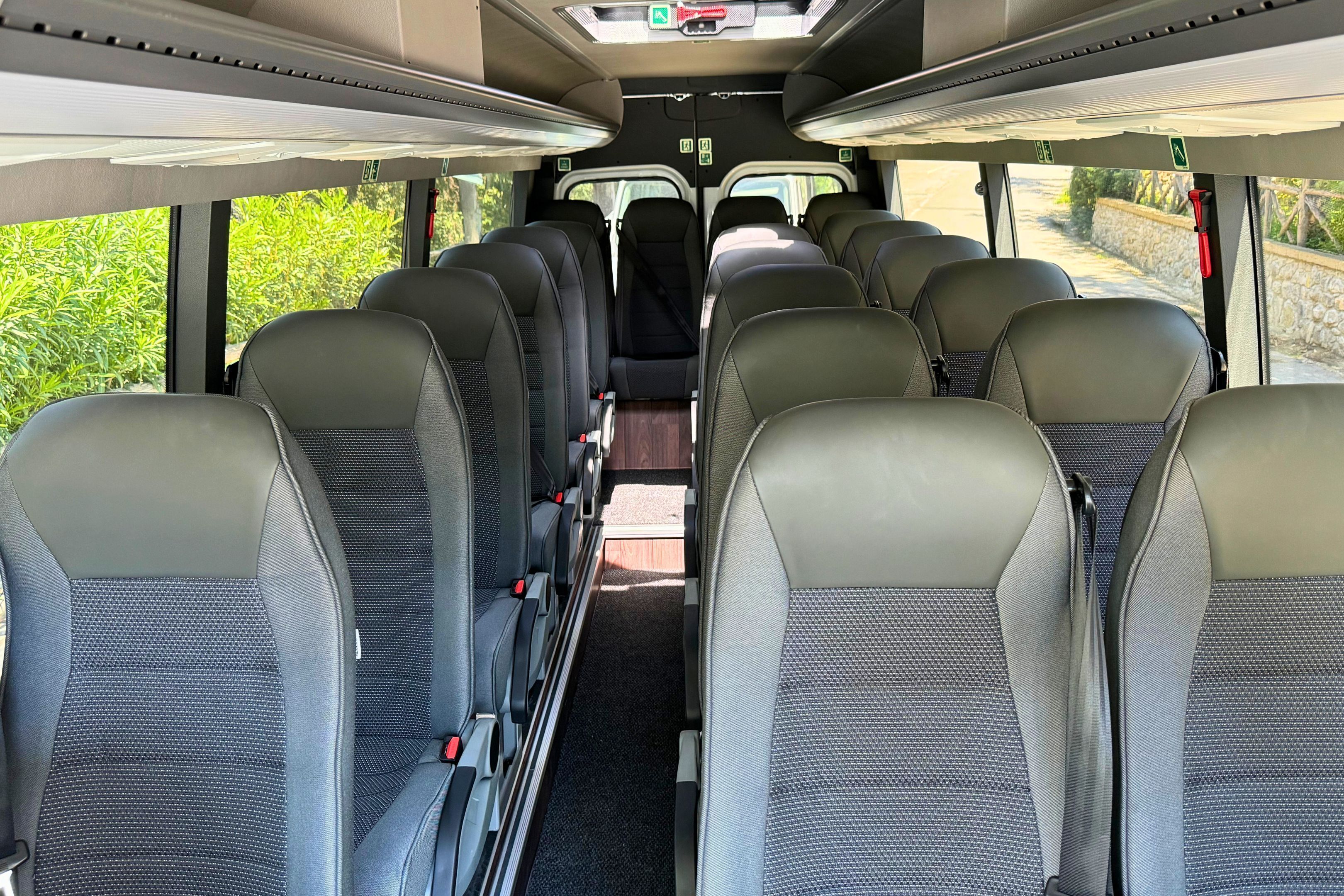 A bus interior with rows of seats, overhead storage compartments, and windows providing a view of greenery outside.