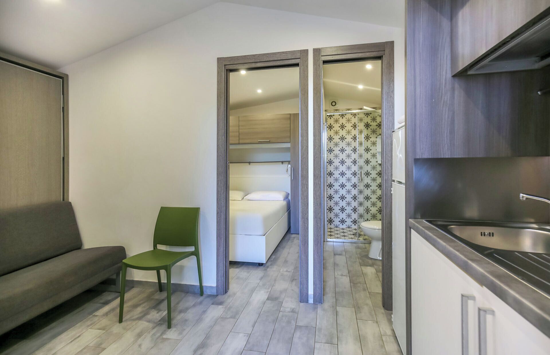 A compact hotel room with a bed, a green chair, a kitchenette, and a shower area.