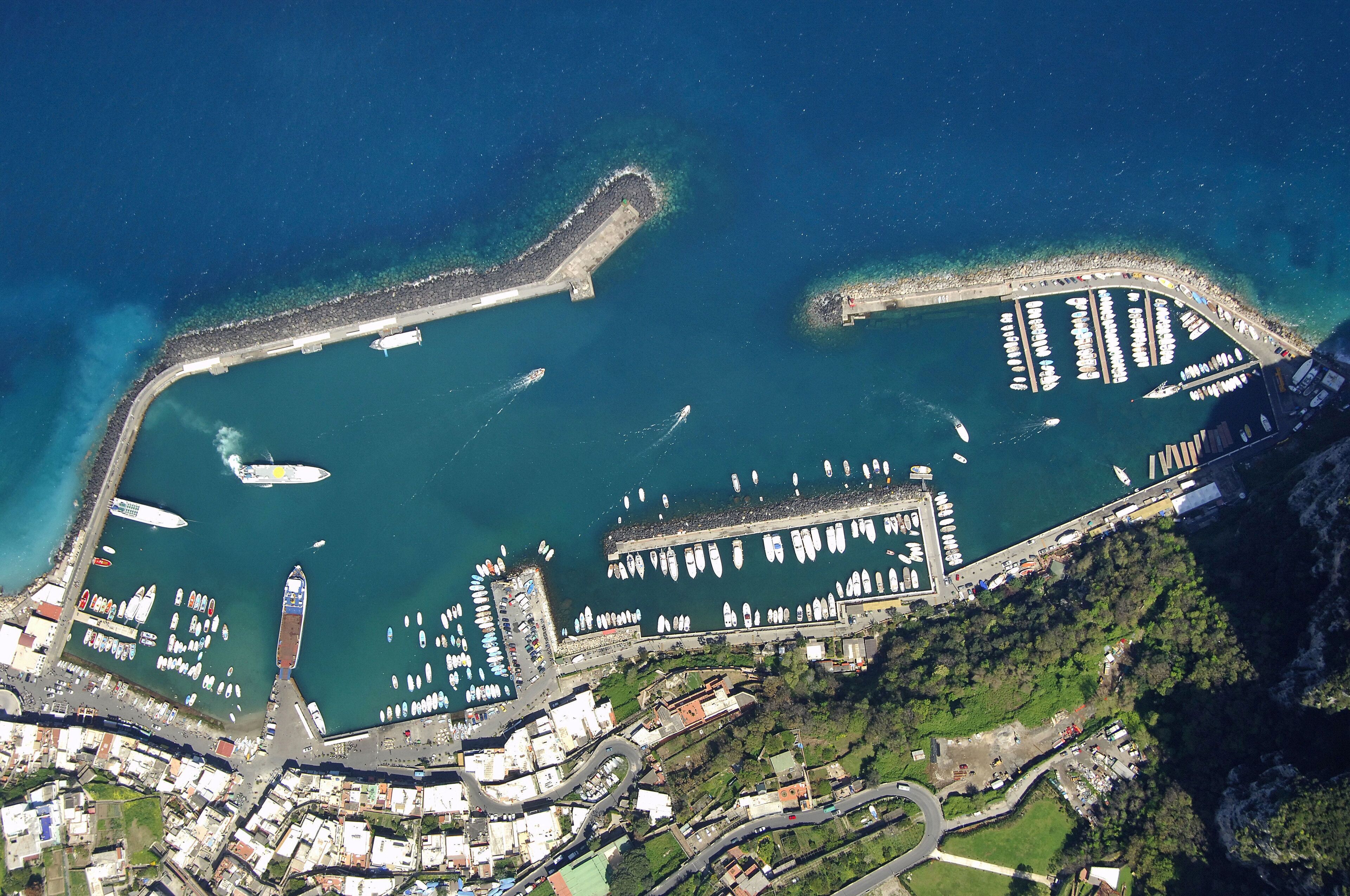 An aerial view of a marina with numerous boats docked, a breakwater, and surrounding buildings.
