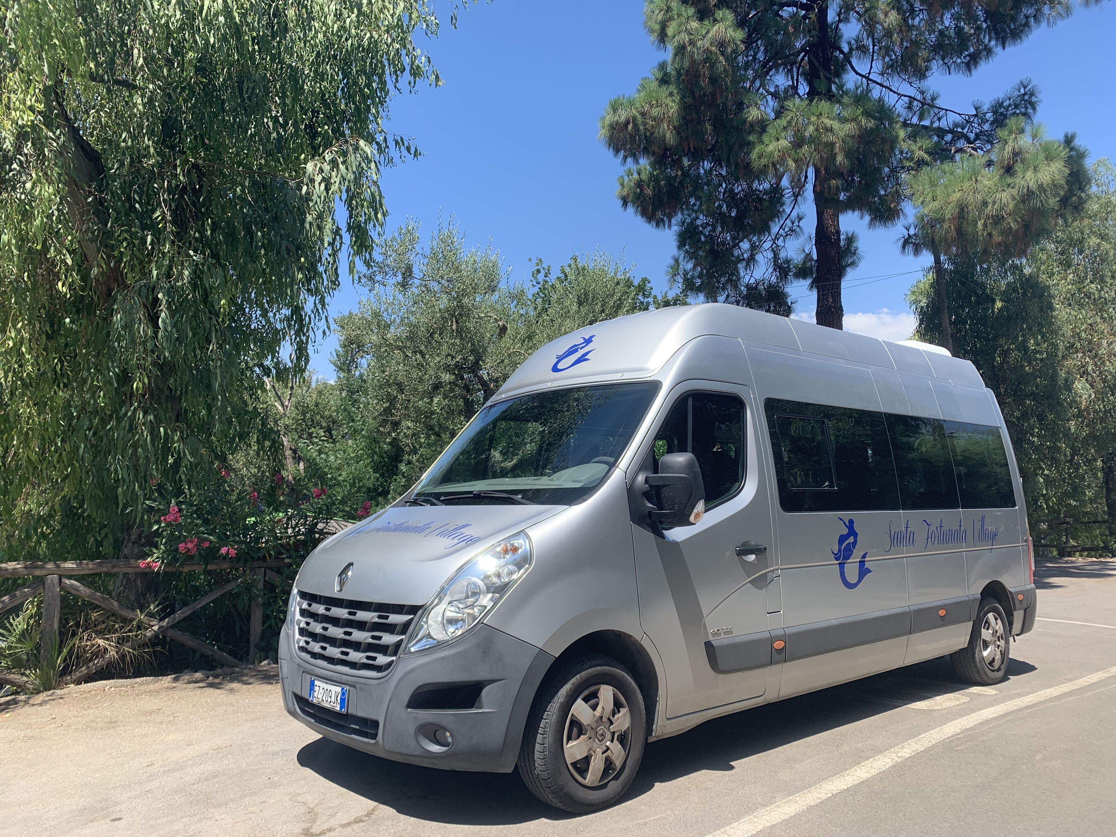 Un fourgon argenté, sur lequel on pouvait lire « Santa Fortuna Village », était garé au bord d’une route bordée d’arbres, avec une clôture en bois en arrière-plan.