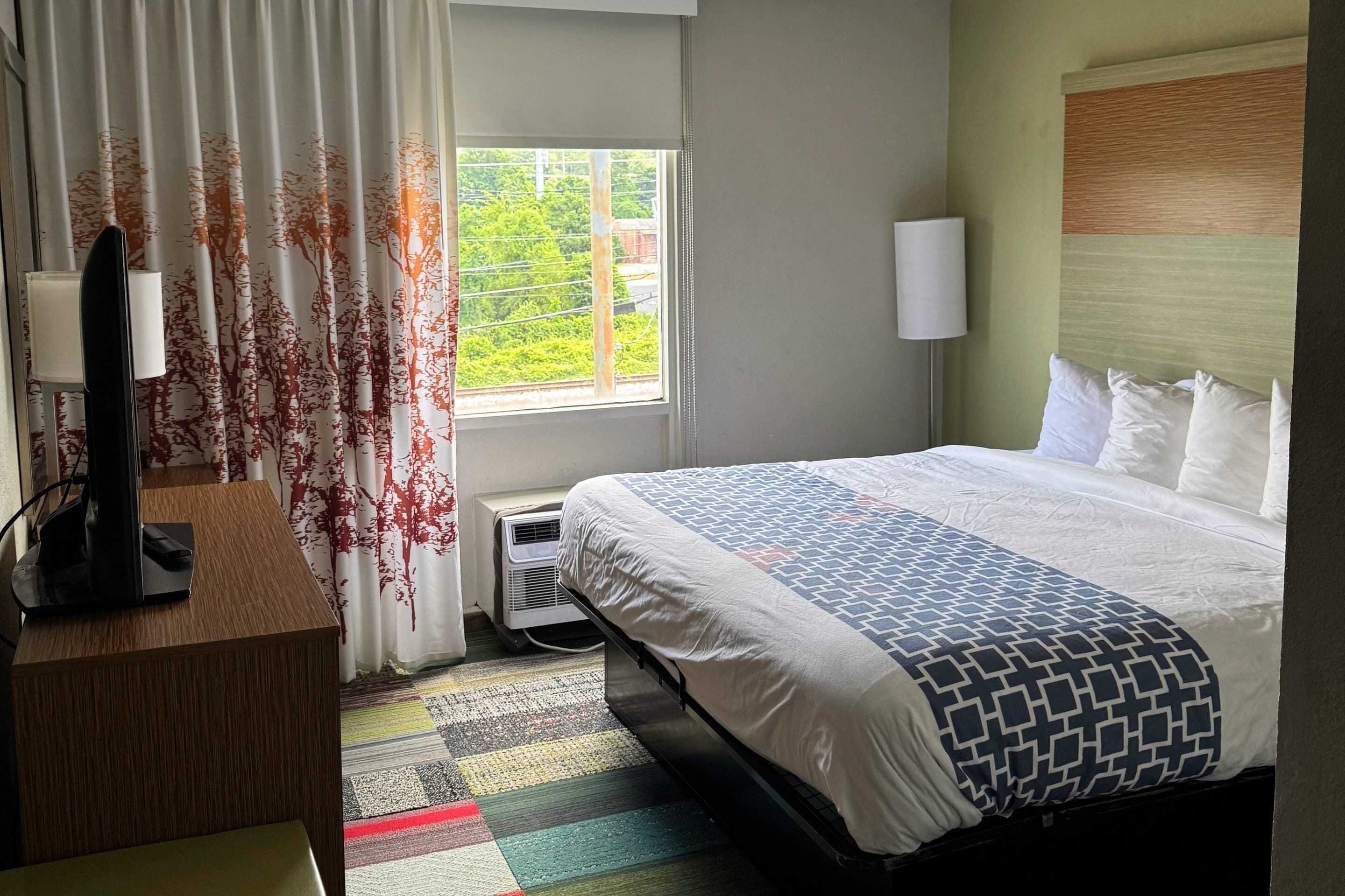 Een hotelkamer met een bed, een bureau met een computer, een raam met gordijnen en een lamp.