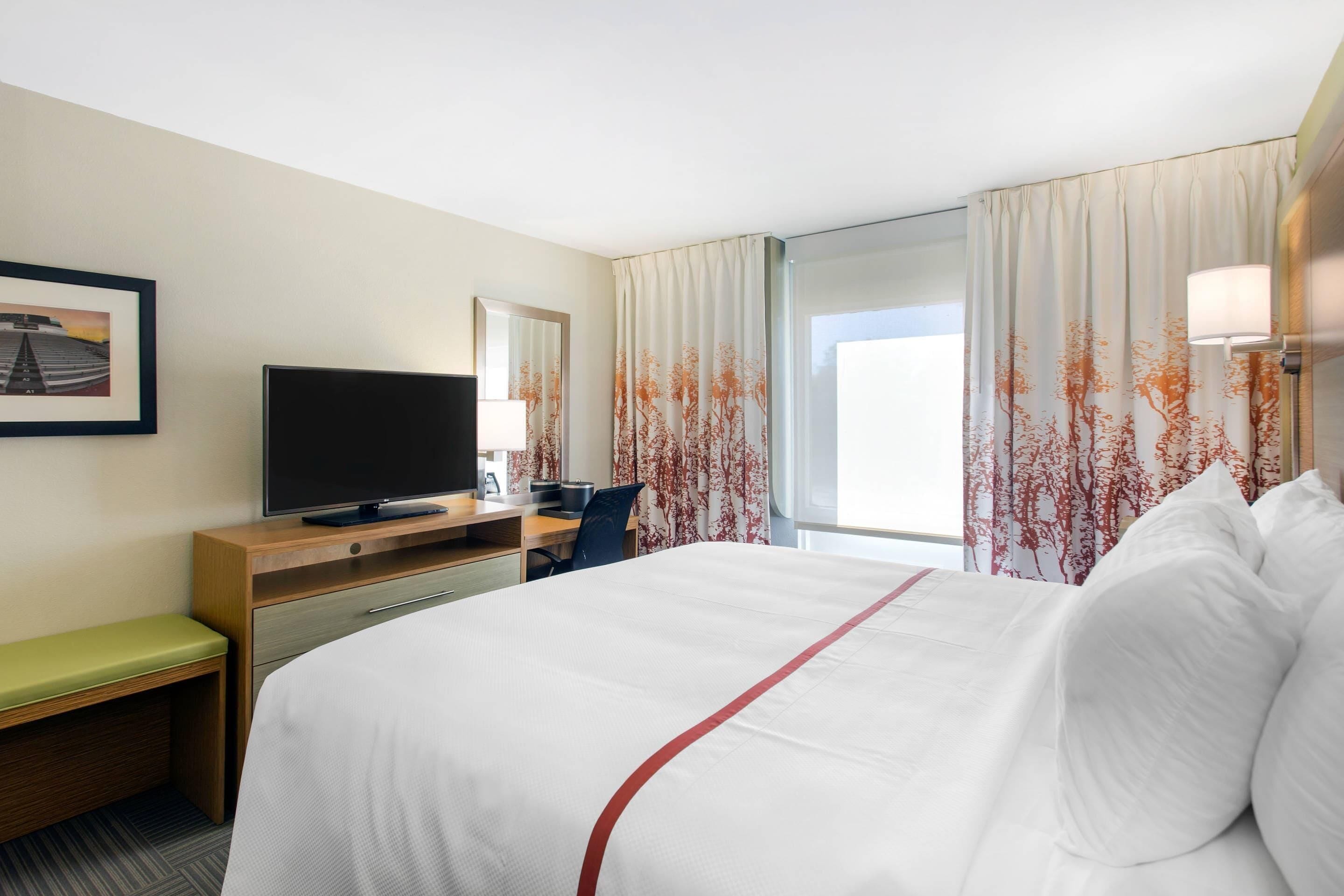 Une chambre d’hôtel avec un lit, un bureau avec une télévision, une chaise et une fenêtre avec des rideaux.