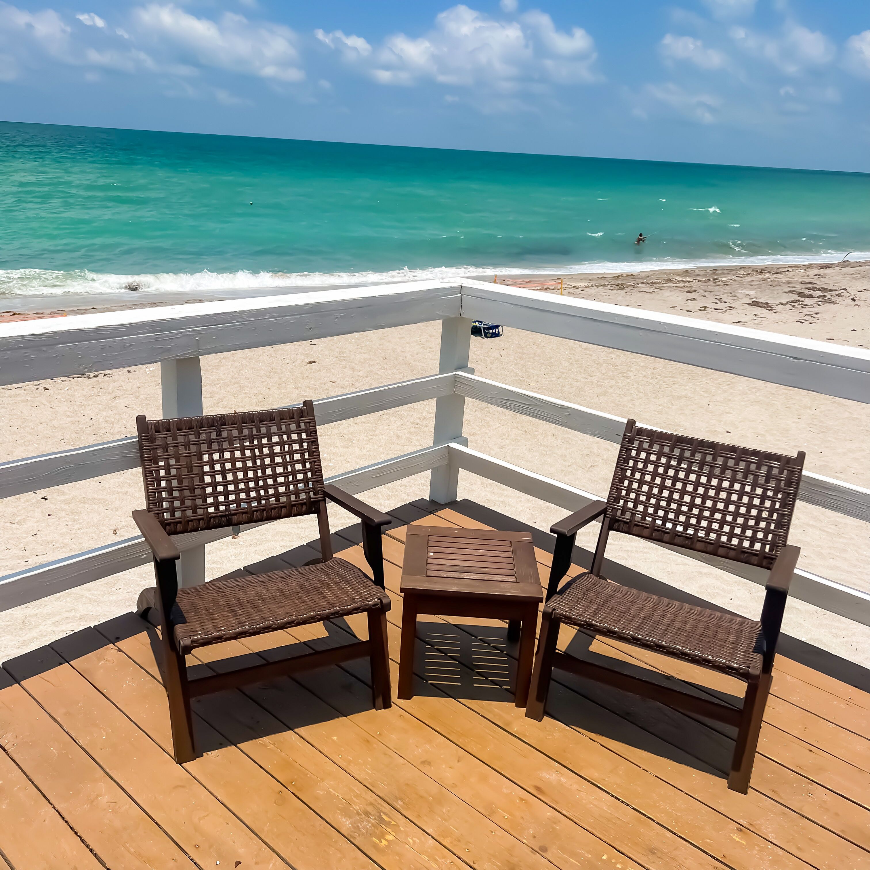A wooden deck with two wicker chairs and a small table, overlooking a beach with turquoise water and a clear sky.