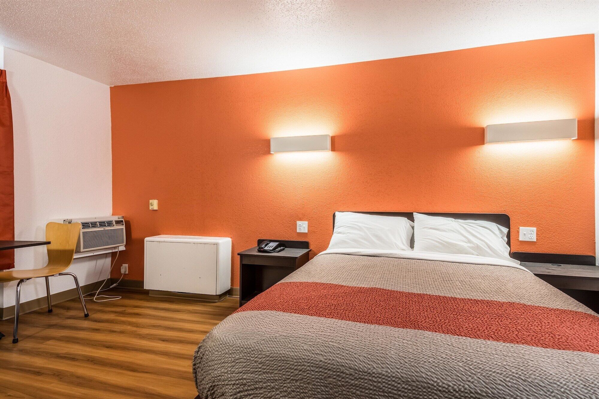 Quarto de hotel com cama, mesas de cabeceira, uma cadeira, aparelho de ar condicionado e duas luminárias fixadas na parede.