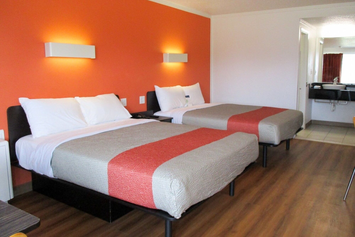 Une chambre d’hôtel avec deux lits, chacun agrémenté de linge de lit blanc et d’un élément décoratif rouge. On y trouve deux tables de chevet avec des lampes, ainsi qu’une petite table avec une chaise dans un coin.