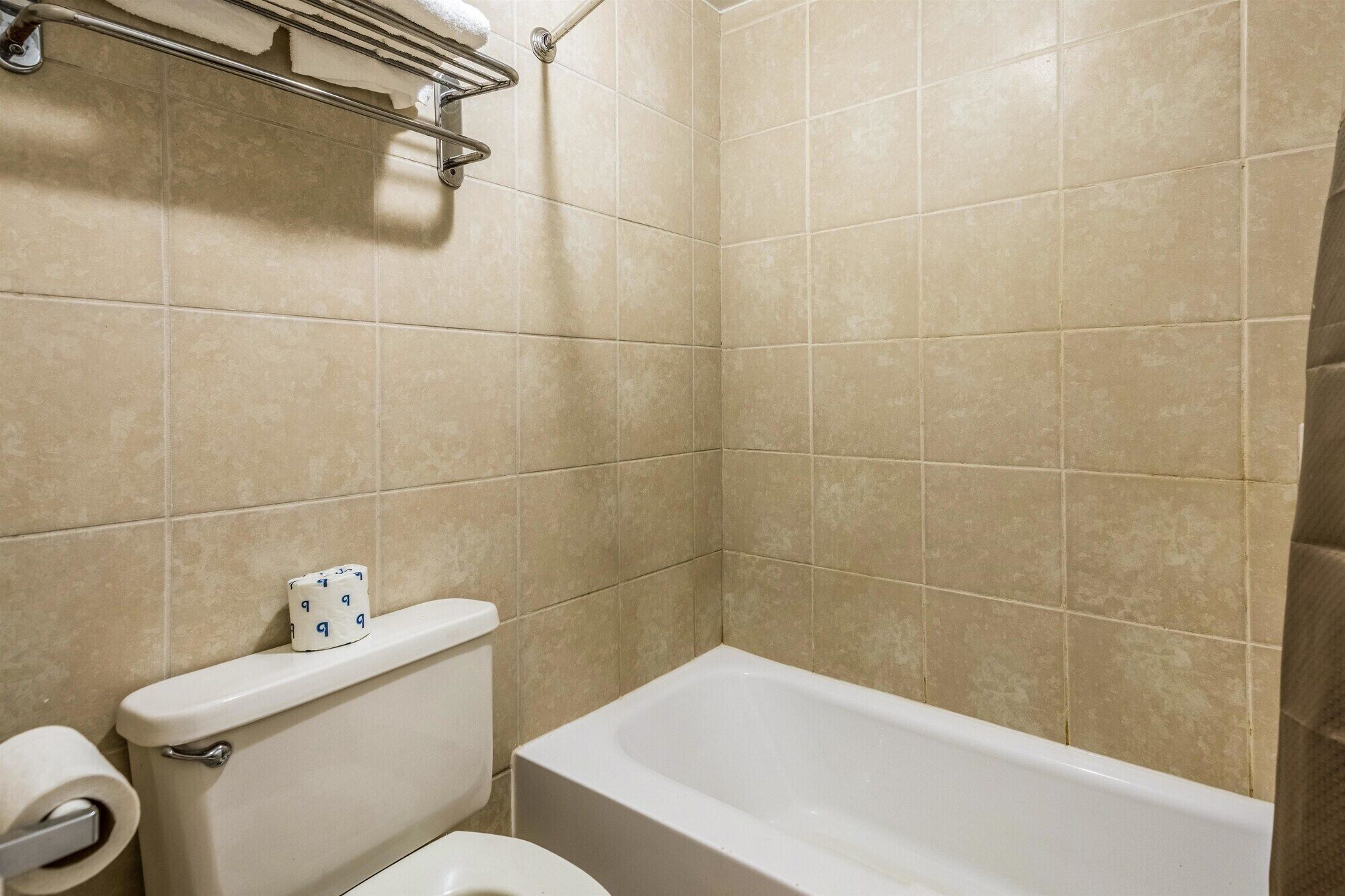 A bathroom with beige tiles, a white toilet, a white bathtub, and a metal towel rack with a white towel.