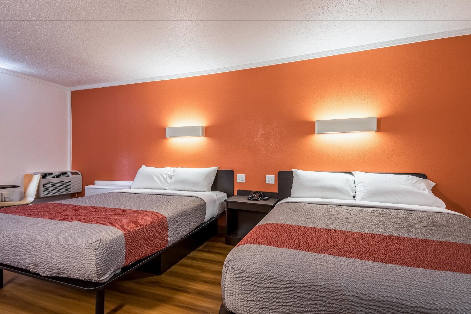 Camera d'albergo con due letti, ciascuno con coperta rossa e grigia, e pavimento in legno. Sono presenti due luci a parete e un condizionatore.