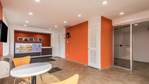 Un moderno vestíbulo de hotel con mostrador de recepción, una mesa redonda blanca con dos sillas y una puerta de cristal que da a una habitación.