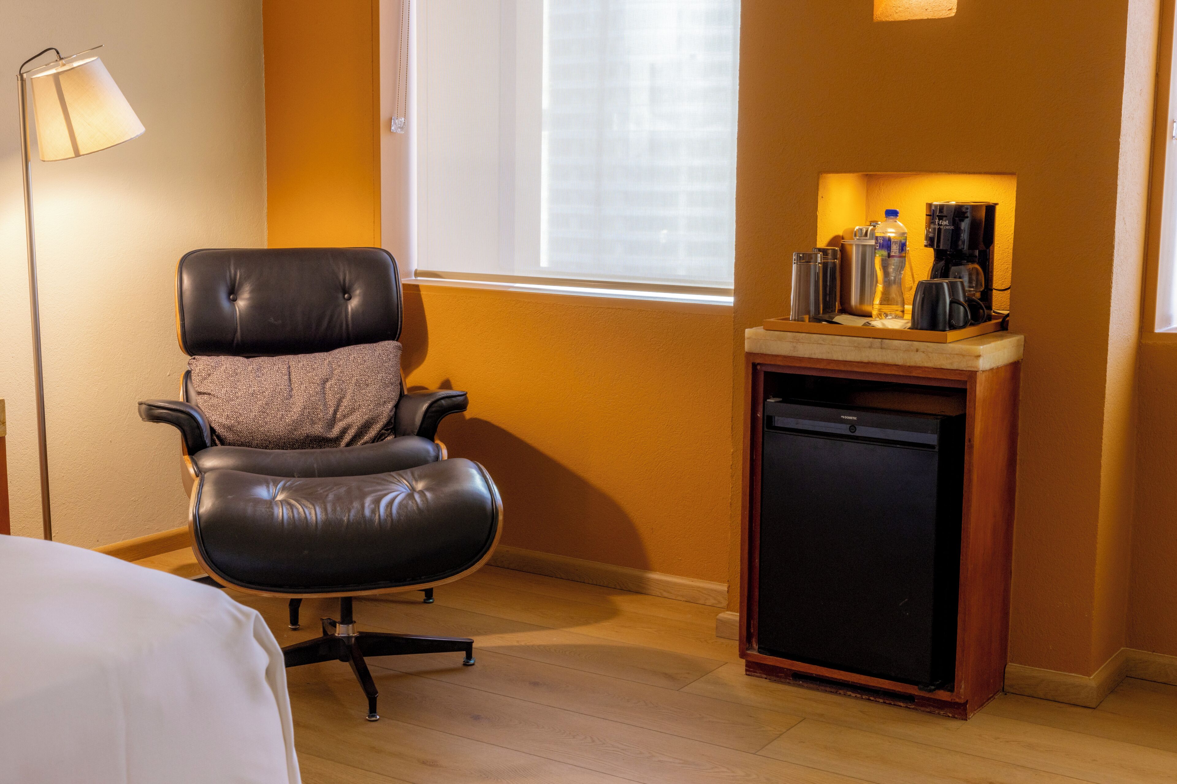 A hotel room with a leather chair, a small wooden table with a coffee maker, a mini fridge, and a bedside table with a lamp.