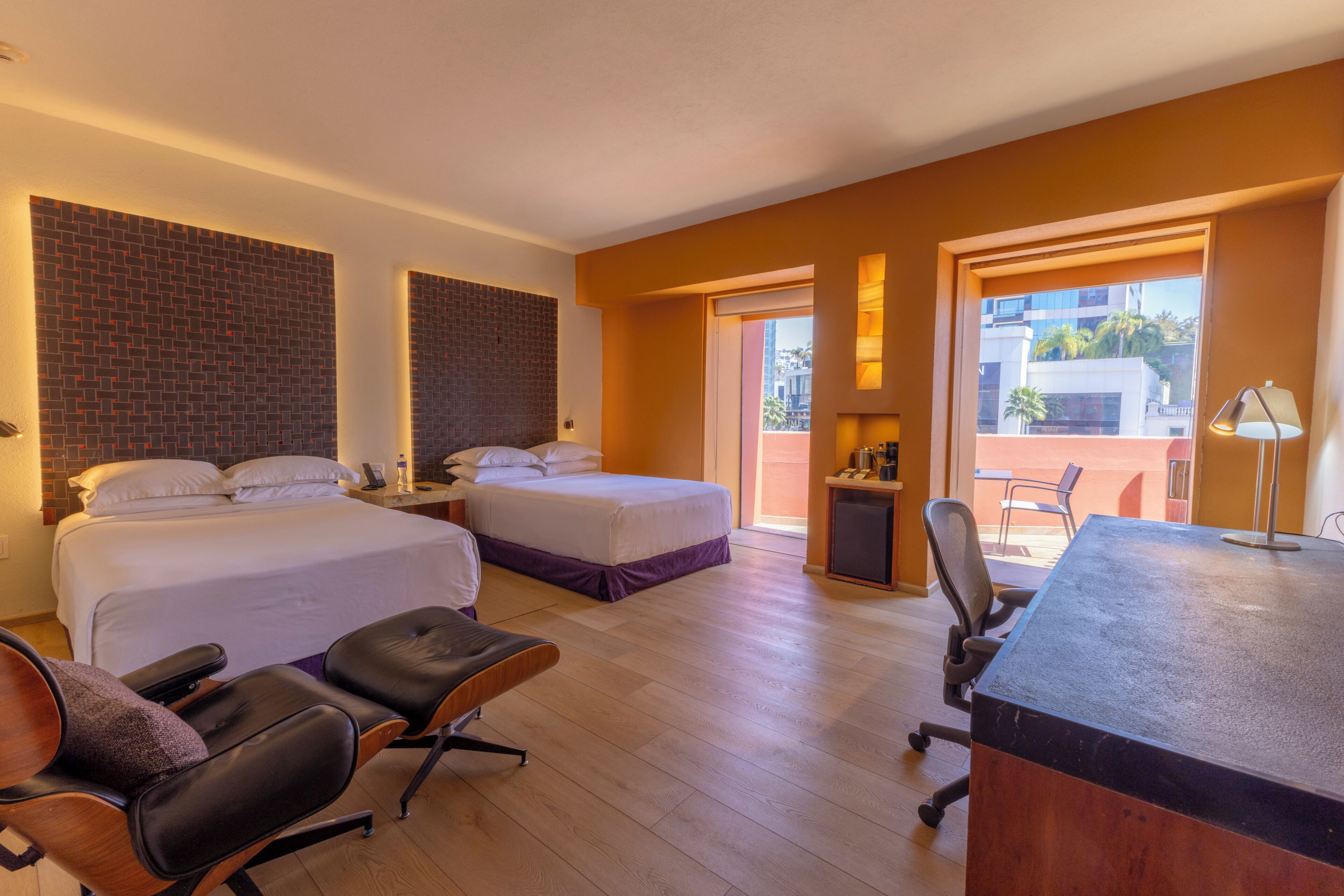 A hotel room with two beds, a desk, a chair, and a view of the outside.