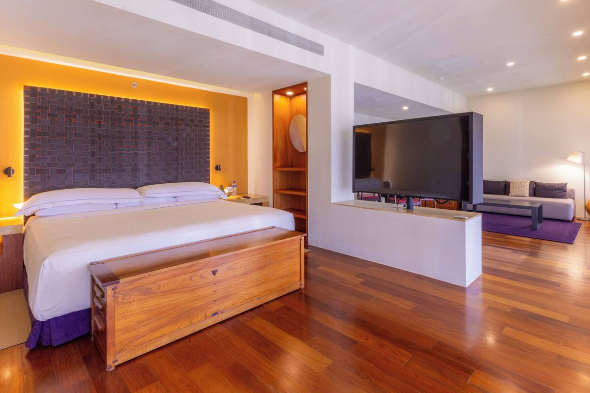 A modern hotel room with a large bed, a flat-screen TV, a wooden bench, a sofa, and a large window.