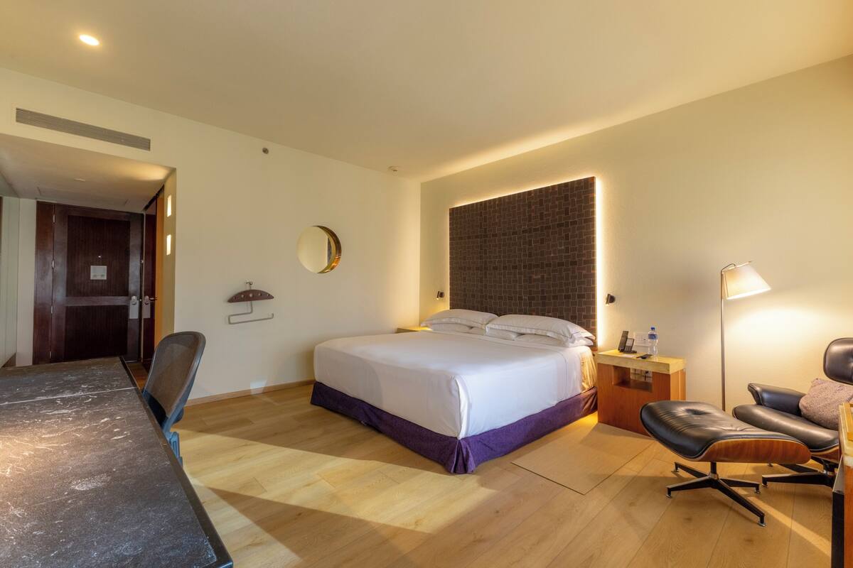 A hotel room with a bed, a desk, a chair, a lamp, and a small table.