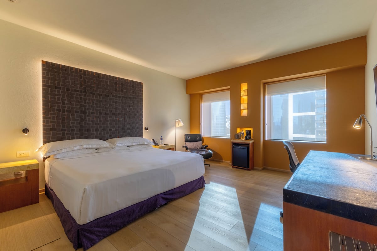 A modern hotel room with a large bed, a desk, a chair, and a small kitchenette.