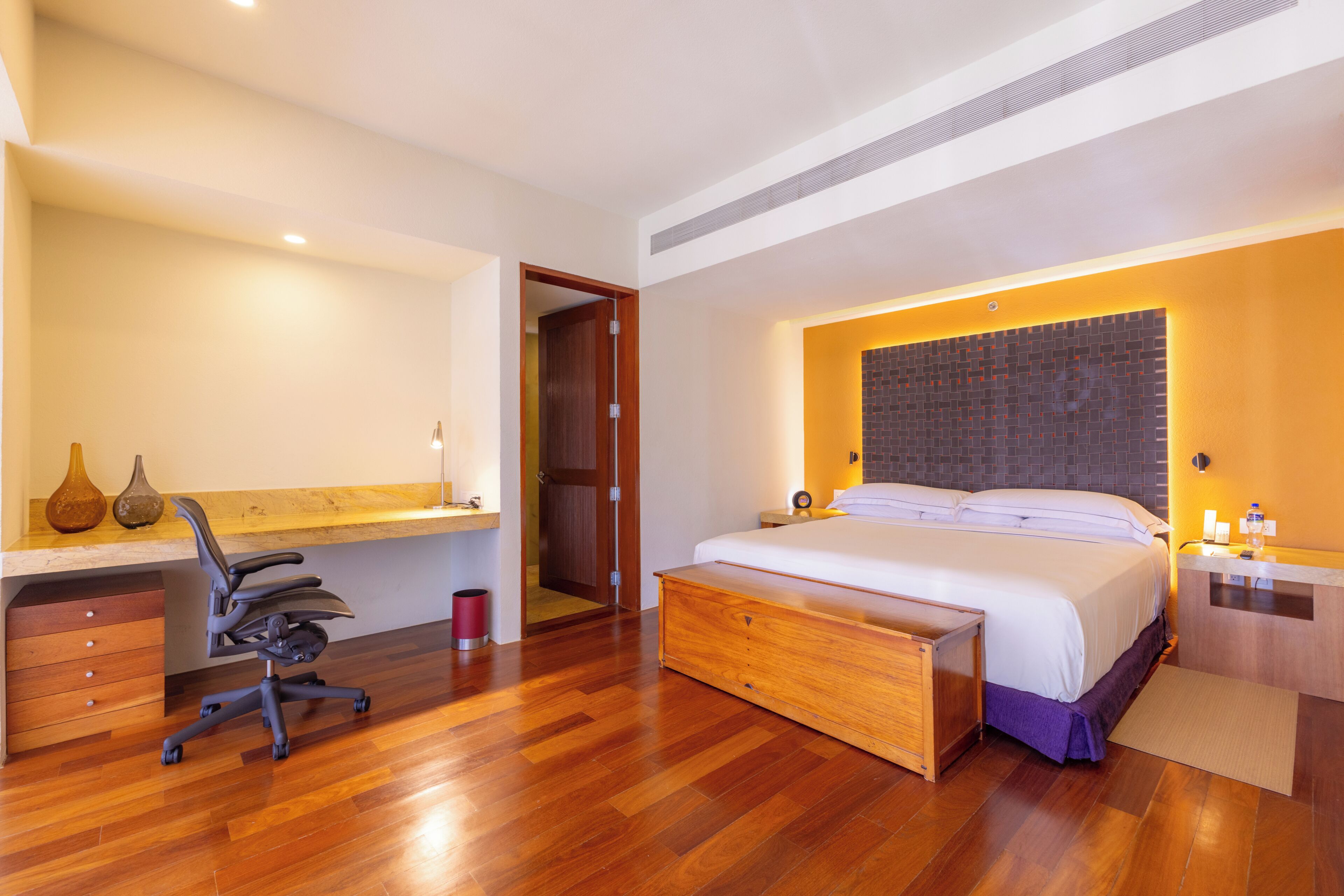 A modern bedroom with a wooden bed, a desk with a chair, a bedside table, and a bathroom visible through an open door.