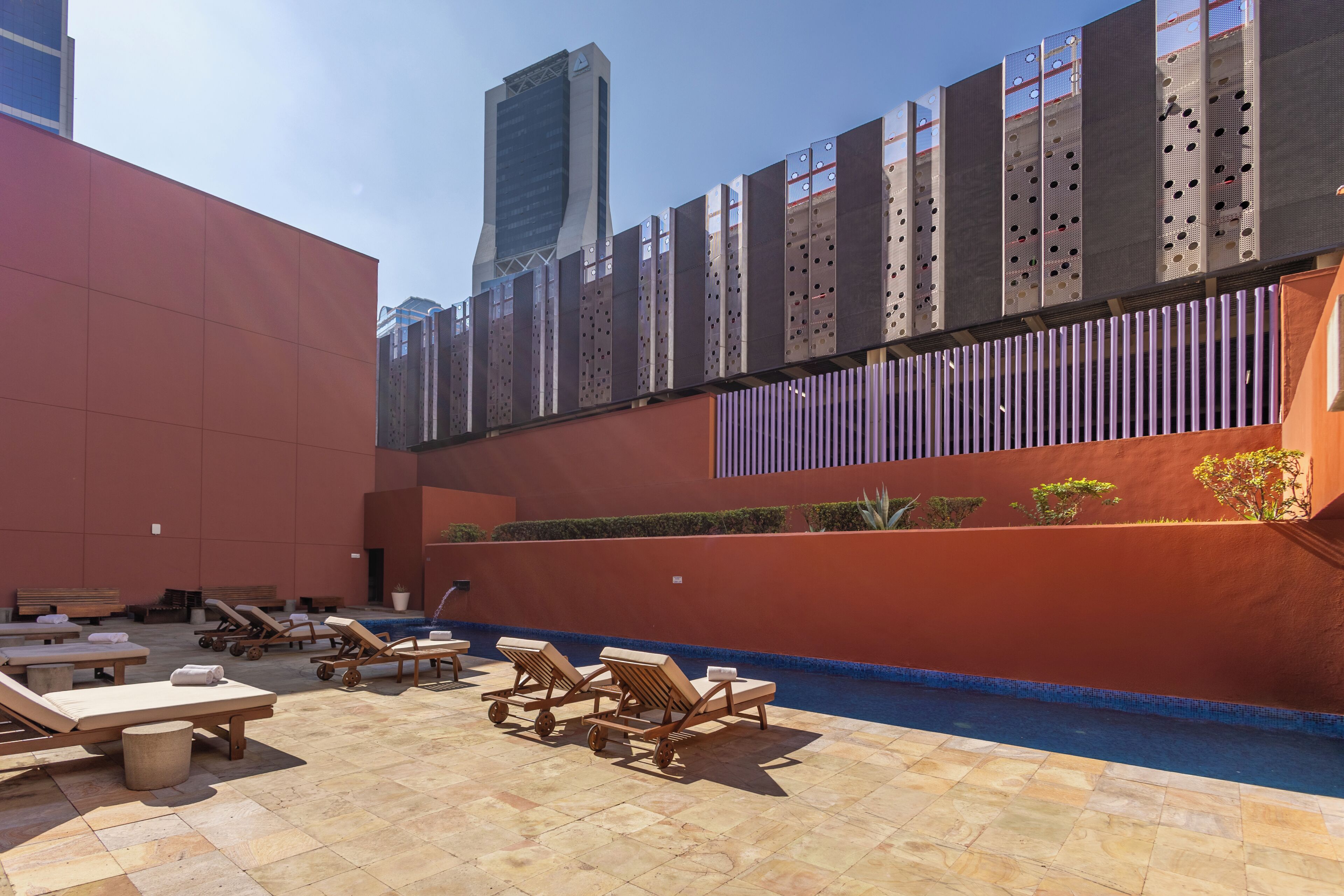 Área de piscina en la azotea con sillones de descanso, una fuente de agua y un edificio moderno al fondo.