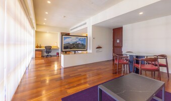 A modern living room with a wooden floor, a flat-screen TV mounted on the wall, a dining table with chairs, and a small coffee table.