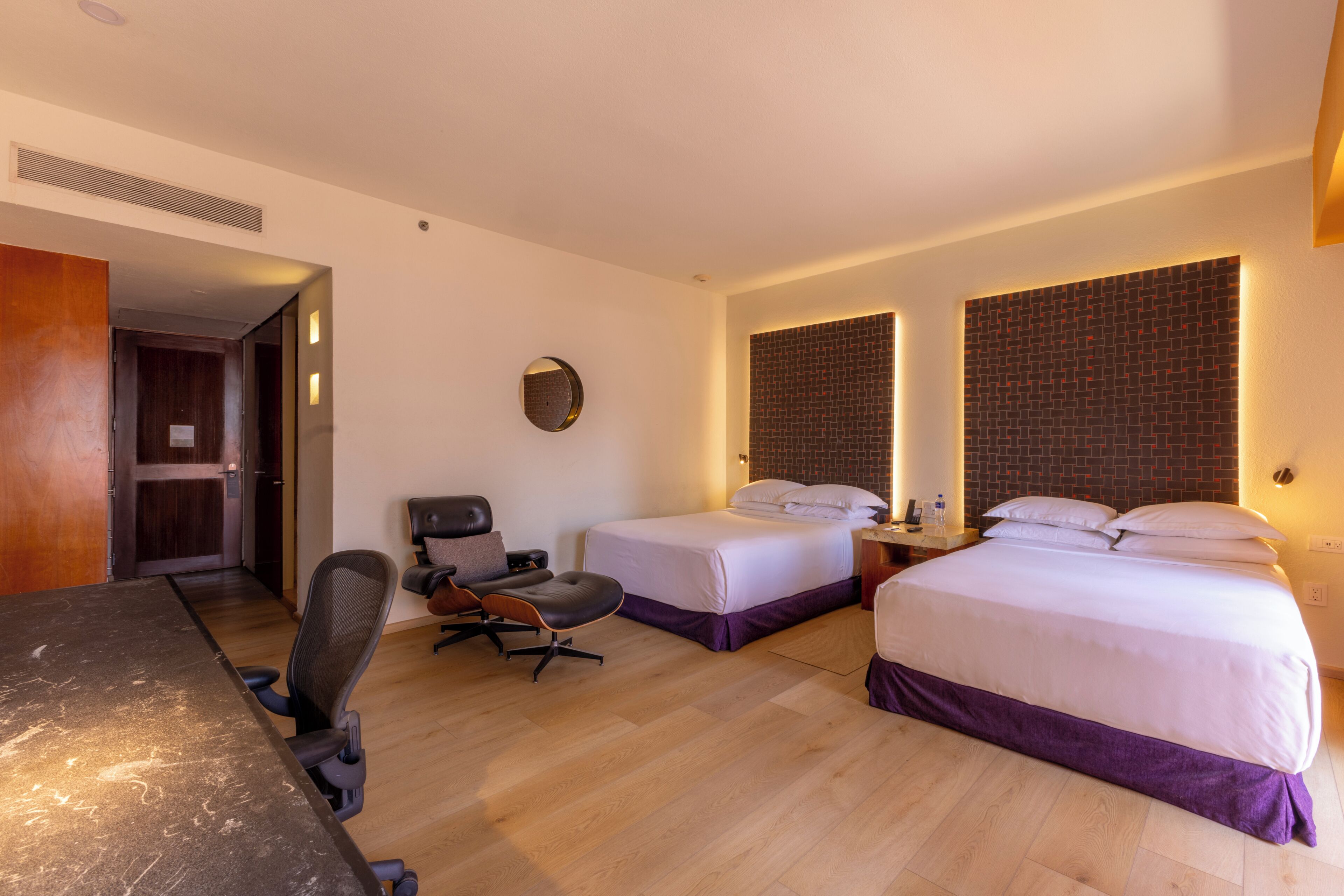 A hotel room with a desk, two beds, a chair, a round mirror, and a brick-patterned wall.