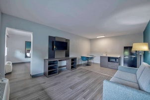 Una sala moderna con un sofá gris, un televisor montado en la pared, una zona de comedor con sillas azules y una kitchenette con microondas y refrigerador.