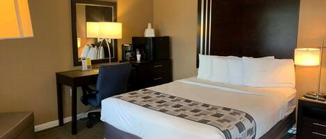 A hotel room with a large bed, a desk with a chair, a dresser, a mirror, and a nightstand with a lamp.