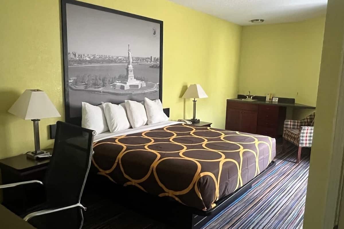 A hotel room with a bed, two bedside tables with lamps, a chair, a desk, a dresser, and a checkered chair.