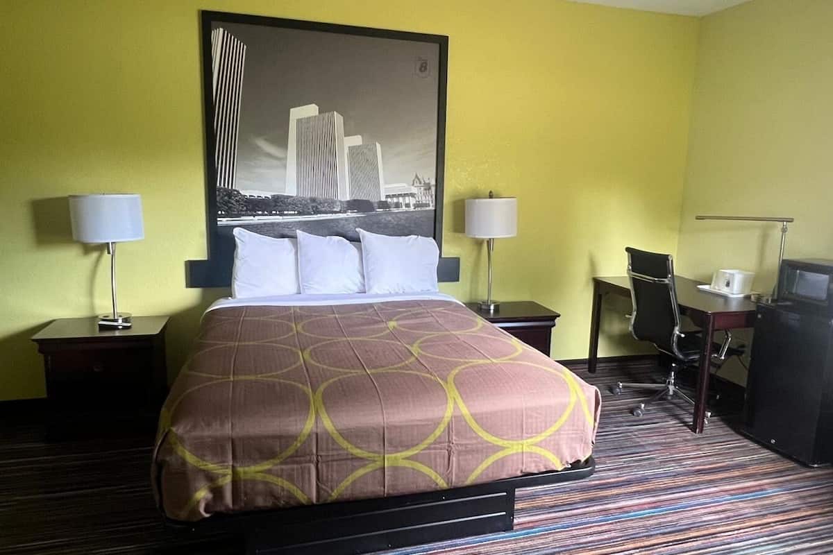 A hotel room with a bed, two bedside tables, a desk, a chair, a TV, and a trash bin.