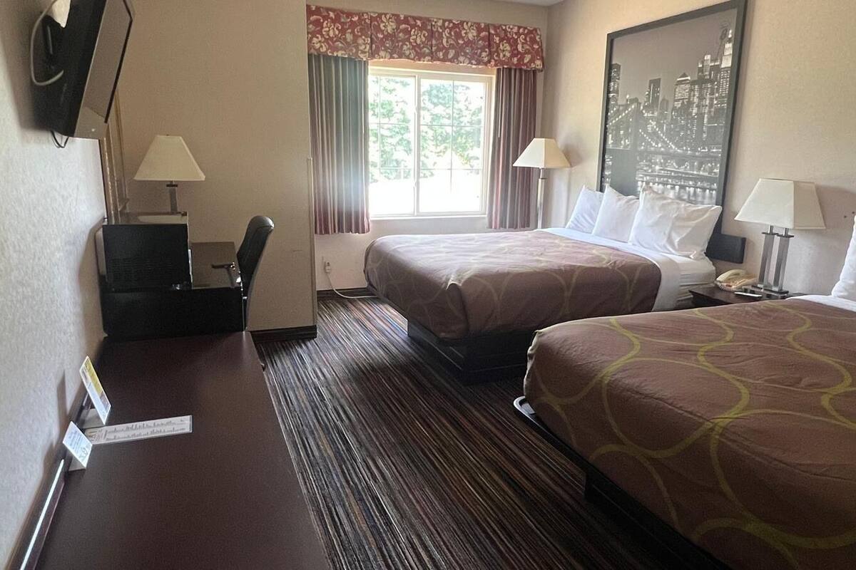 A hotel room with two beds, a desk with a chair, a window with curtains, and a cityscape painting on the wall.