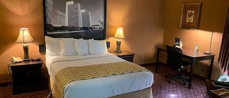 Habitación de hotel con una cama grande, dos mesitas de noche con lámparas, un escritorio con una silla y un cuadro de paisaje urbano en la pared.