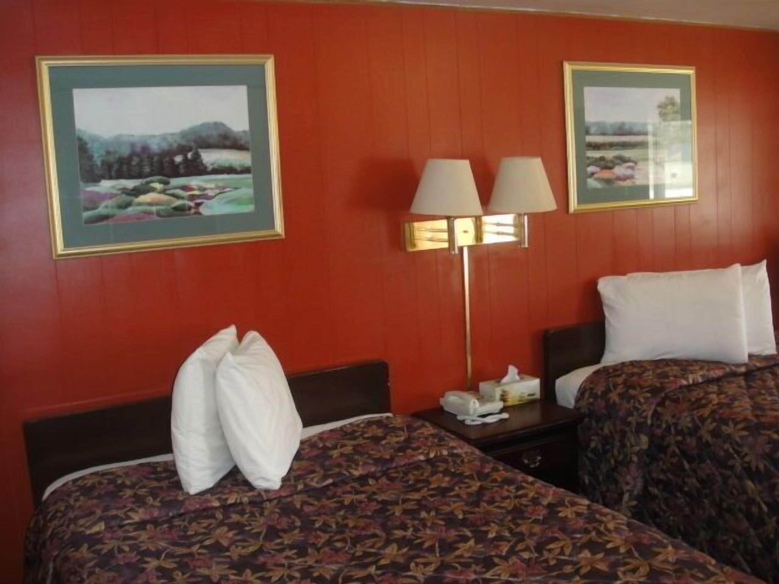 A hotel room with two beds, each with a floral-patterned bedspread, a nightstand with a lamp, and two framed landscape paintings on the walls.
