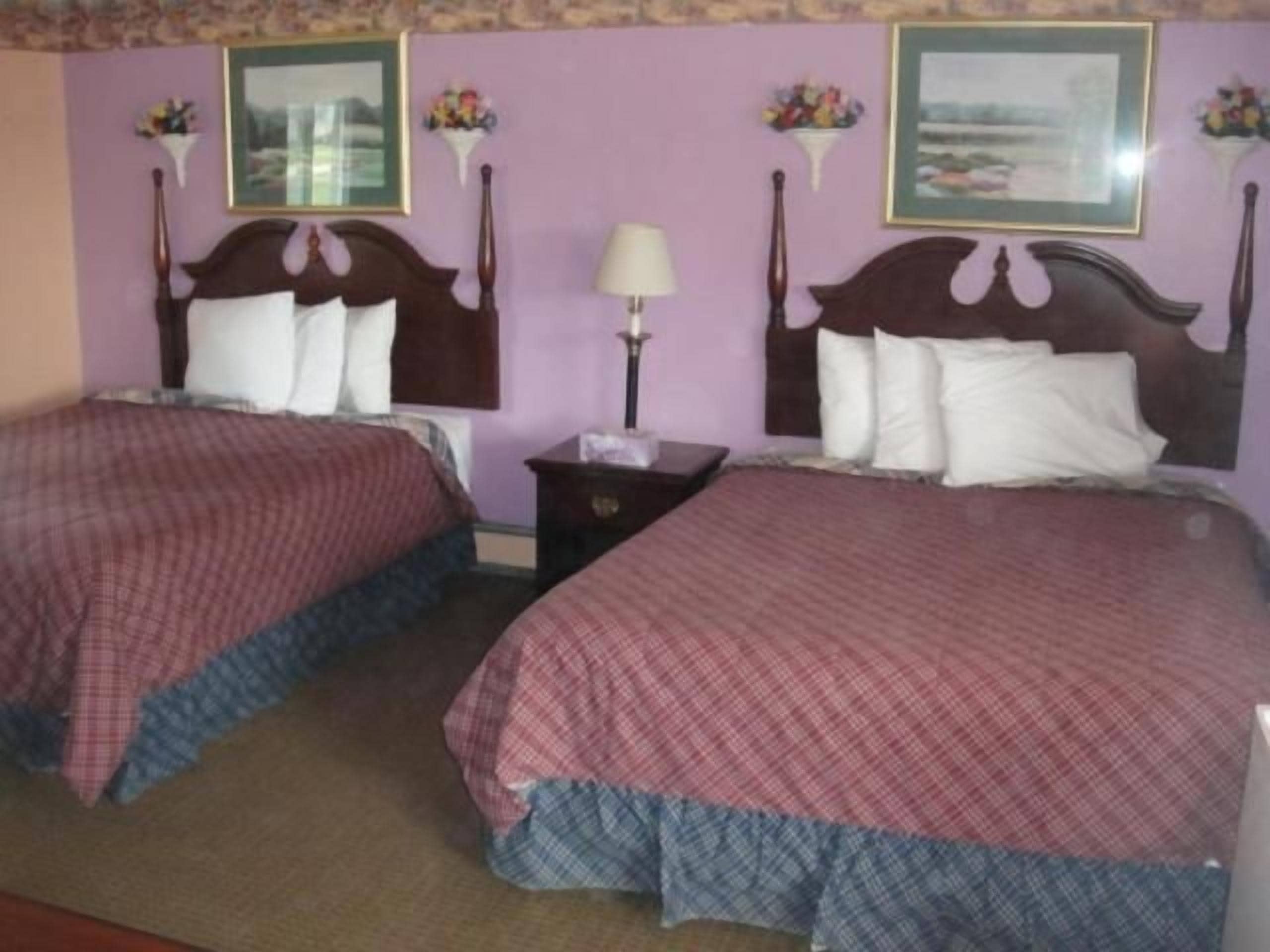 A hotel room with two beds, each with a wooden headboard and a checkered bedspread. There are wall-mounted lamps and a small table between the beds.