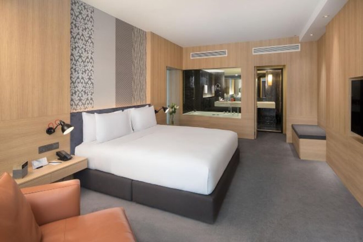 A modern hotel room with a large bed, a desk with a lamp, a chair, and a TV.