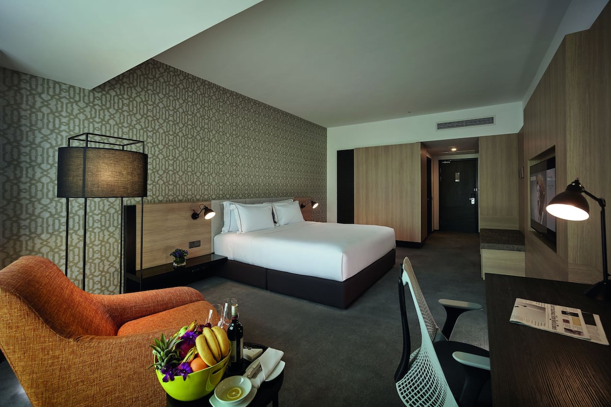 Habitación de hotel moderna con una cama grande, un escritorio, una silla y un sofá. Hay una mesita con un frutero y una botella.