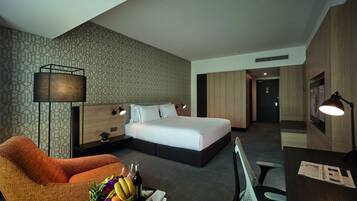 Een moderne hotelkamer met een groot bed, een bureau, een stoel en een bank. Er staat een klein tafeltje met een fruitschaal en een fles.