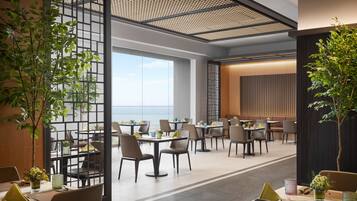 A modern dining area with tables and chairs, large windows, and indoor plants.