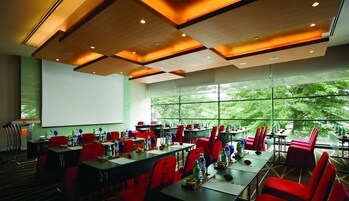 A conference room with tables and red chairs, large windows with a view of trees, and a projector screen.