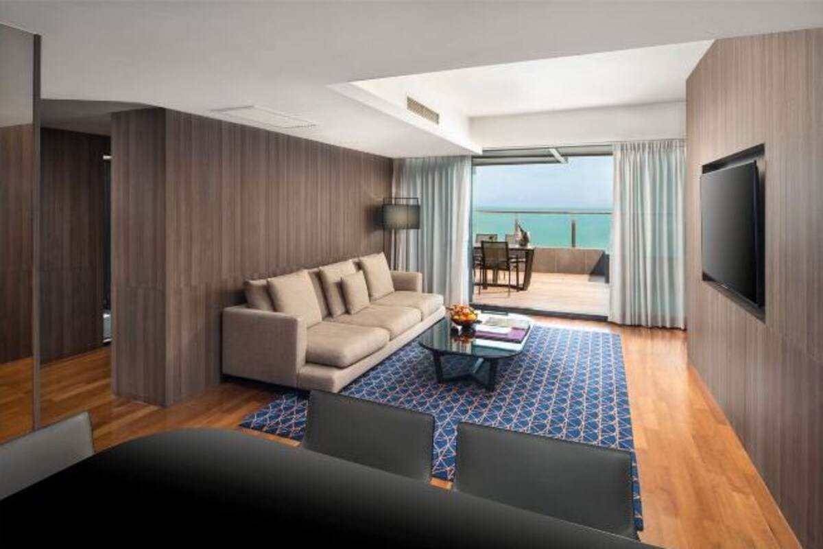 A modern hotel room with a sofa, a coffee table, and a TV. There is a balcony with a view of the sea.