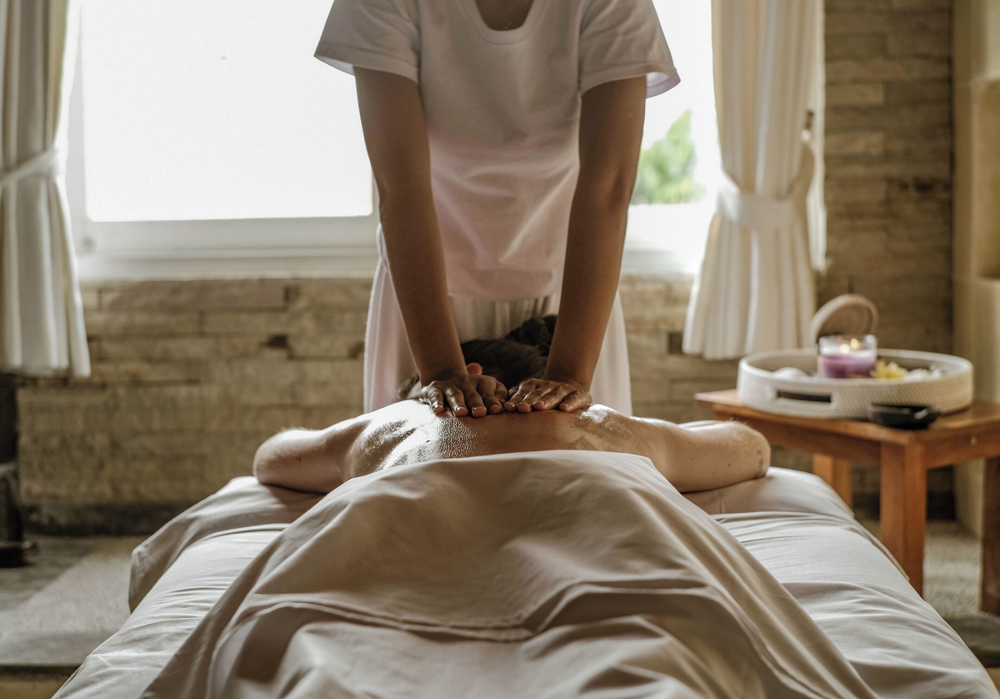A massage therapy session in a room with a brick wall, a wooden table with a candle, and a window with curtains.
