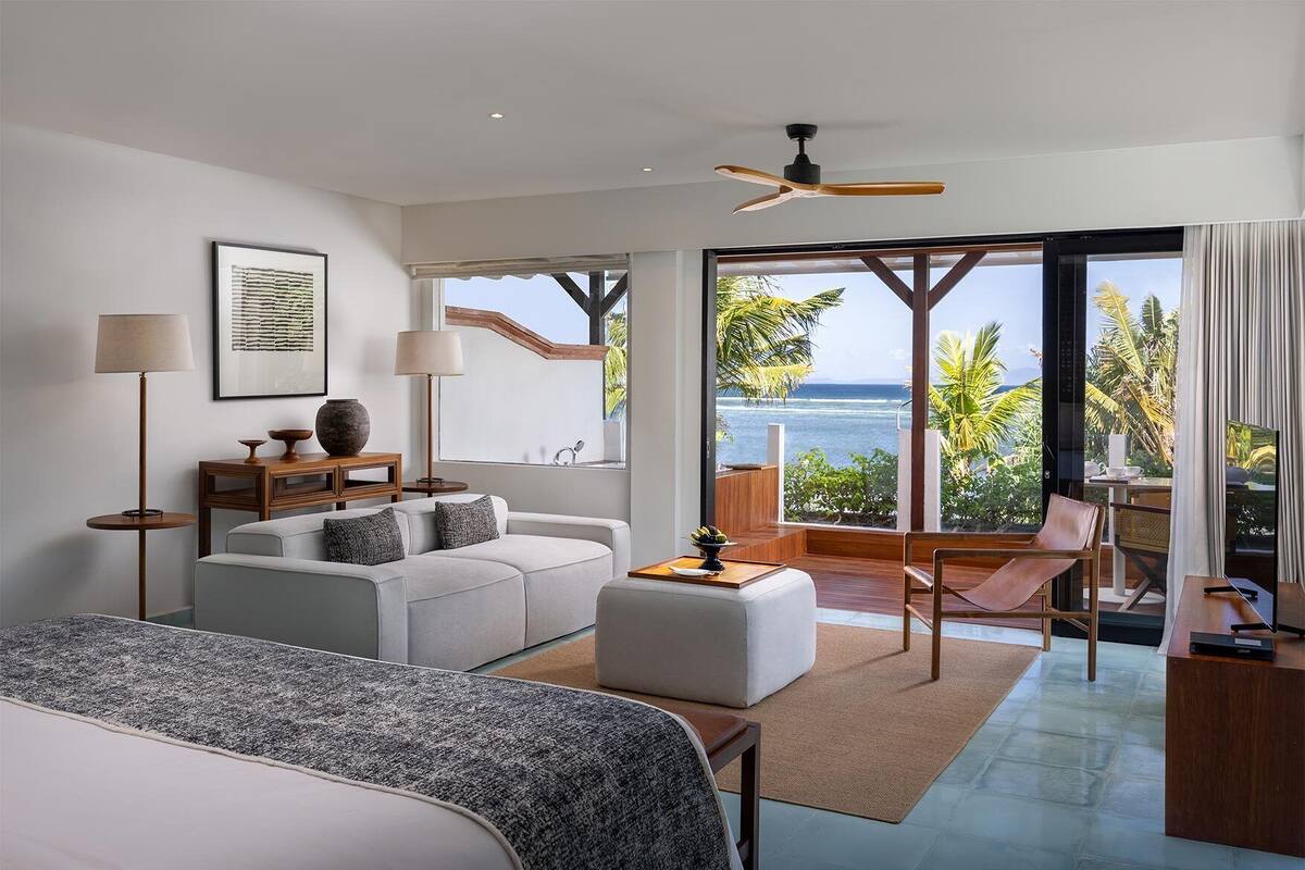 A modern hotel room with a large bed, a sofa, a coffee table, and a balcony with a view of the ocean.