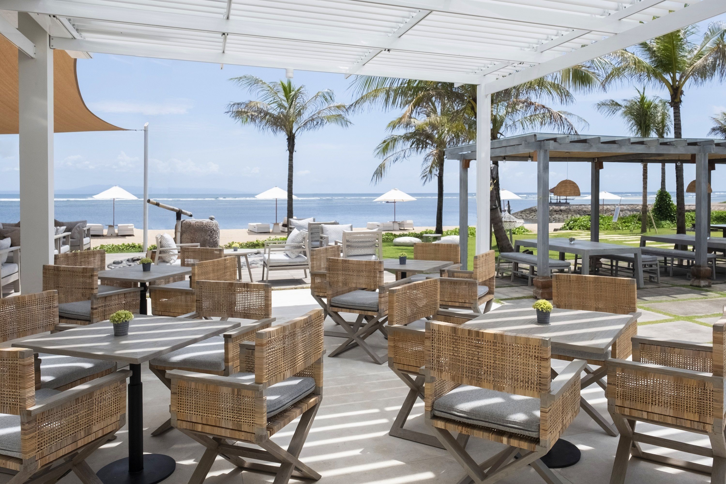 An outdoor seating area with wicker furniture and umbrellas, overlooking a beach with palm trees and the ocean.
