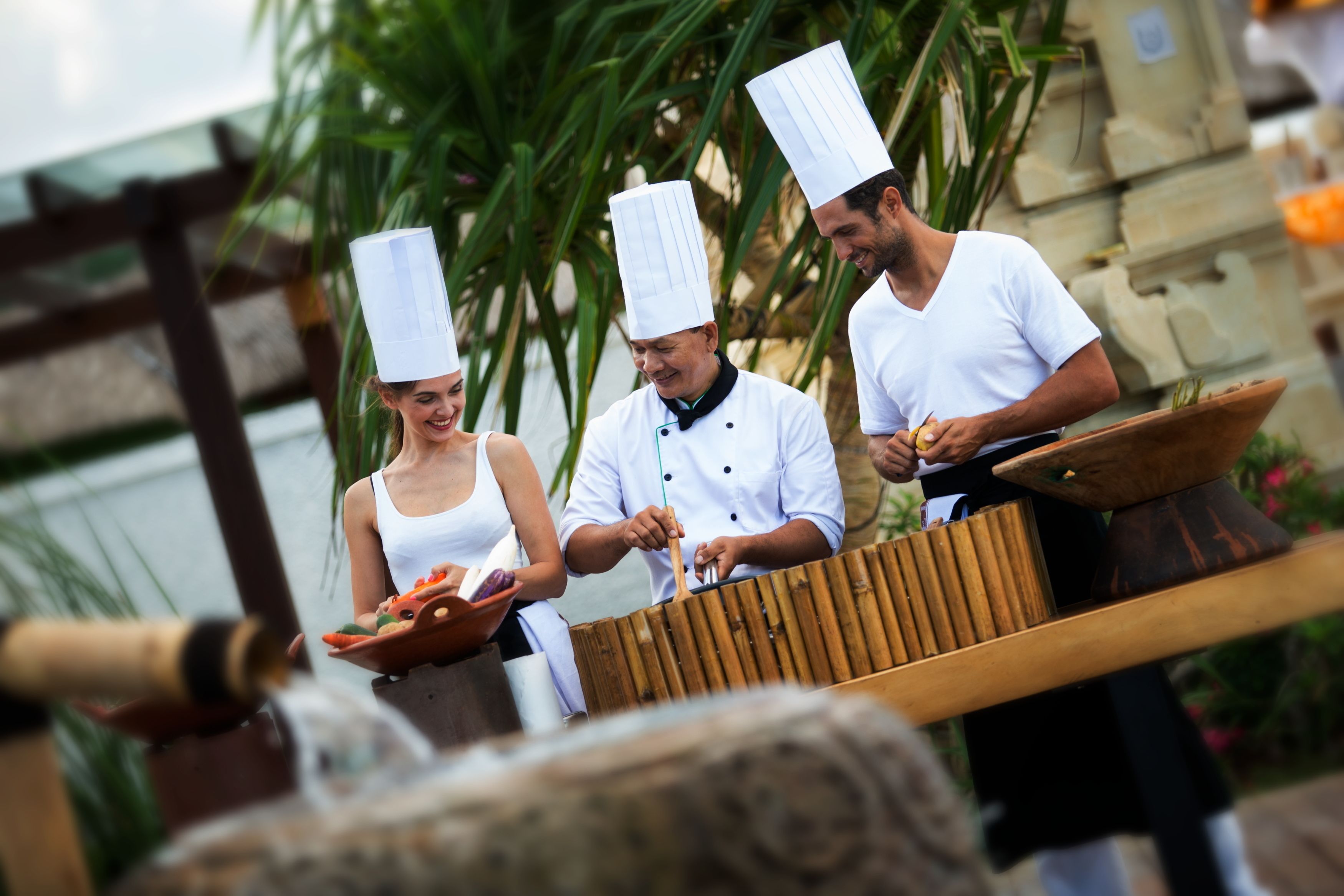 Cocineros con uniformes blancos y sombreros altos preparando comida al aire libre, con una estructura de madera y follaje de fondo.