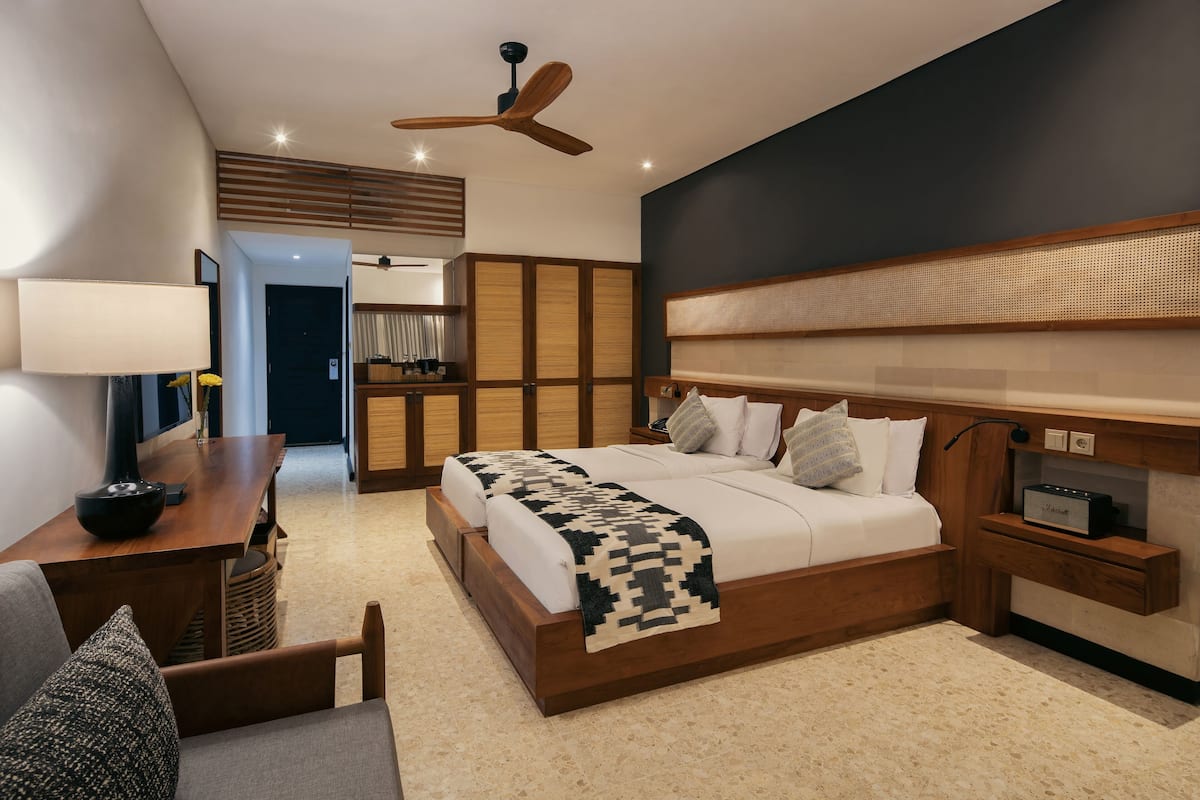 A modern hotel room with a large bed, a desk with a lamp, a ceiling fan, and a small bedside table with a radio.