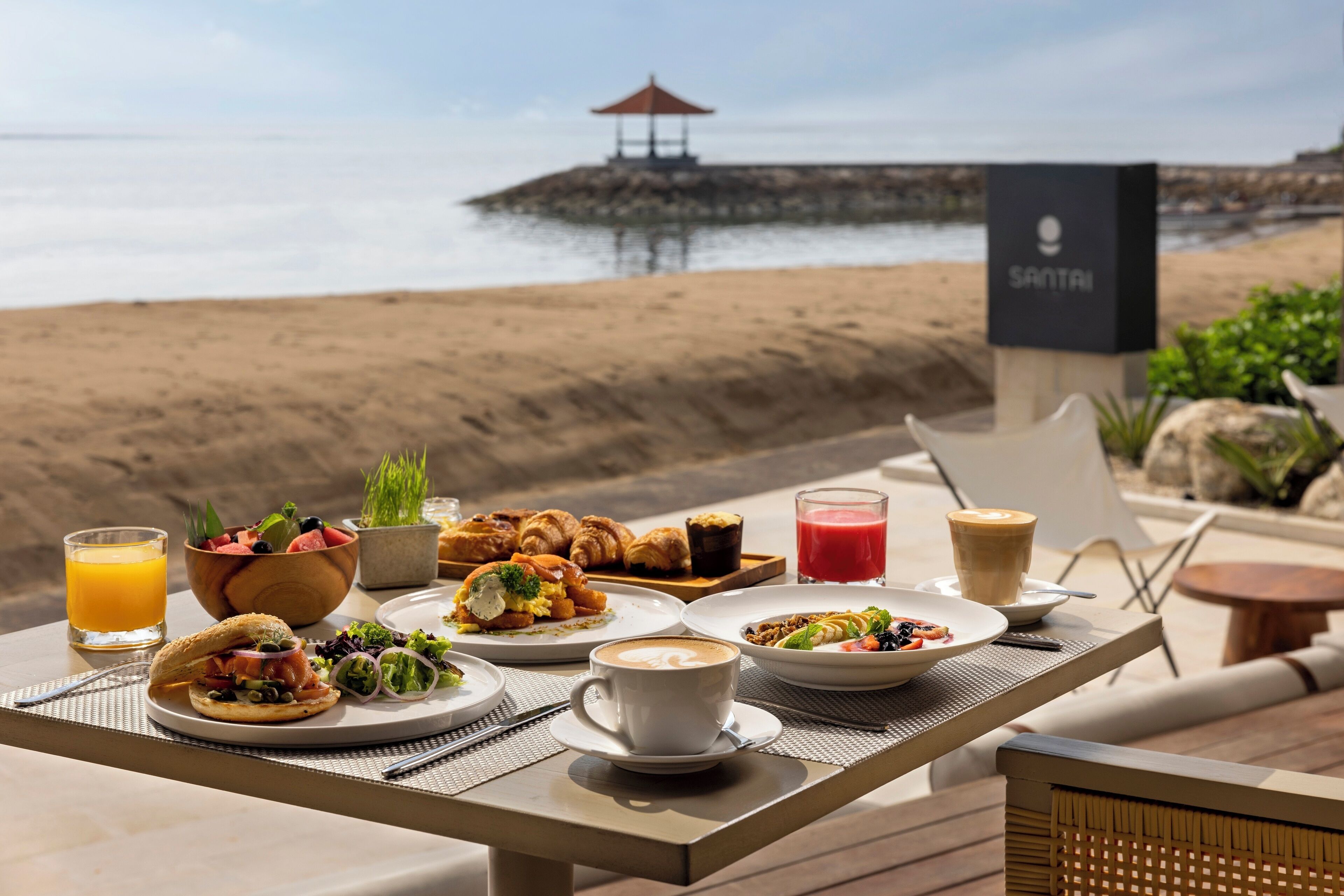 A breakfast table with various dishes, a glass of orange juice, and a view of the beach and ocean.