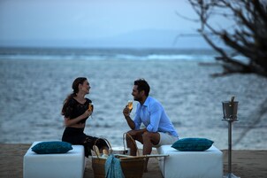 Two people sitting on white chairs by the sea, holding drinks. There is a basket and a lamp on the table.