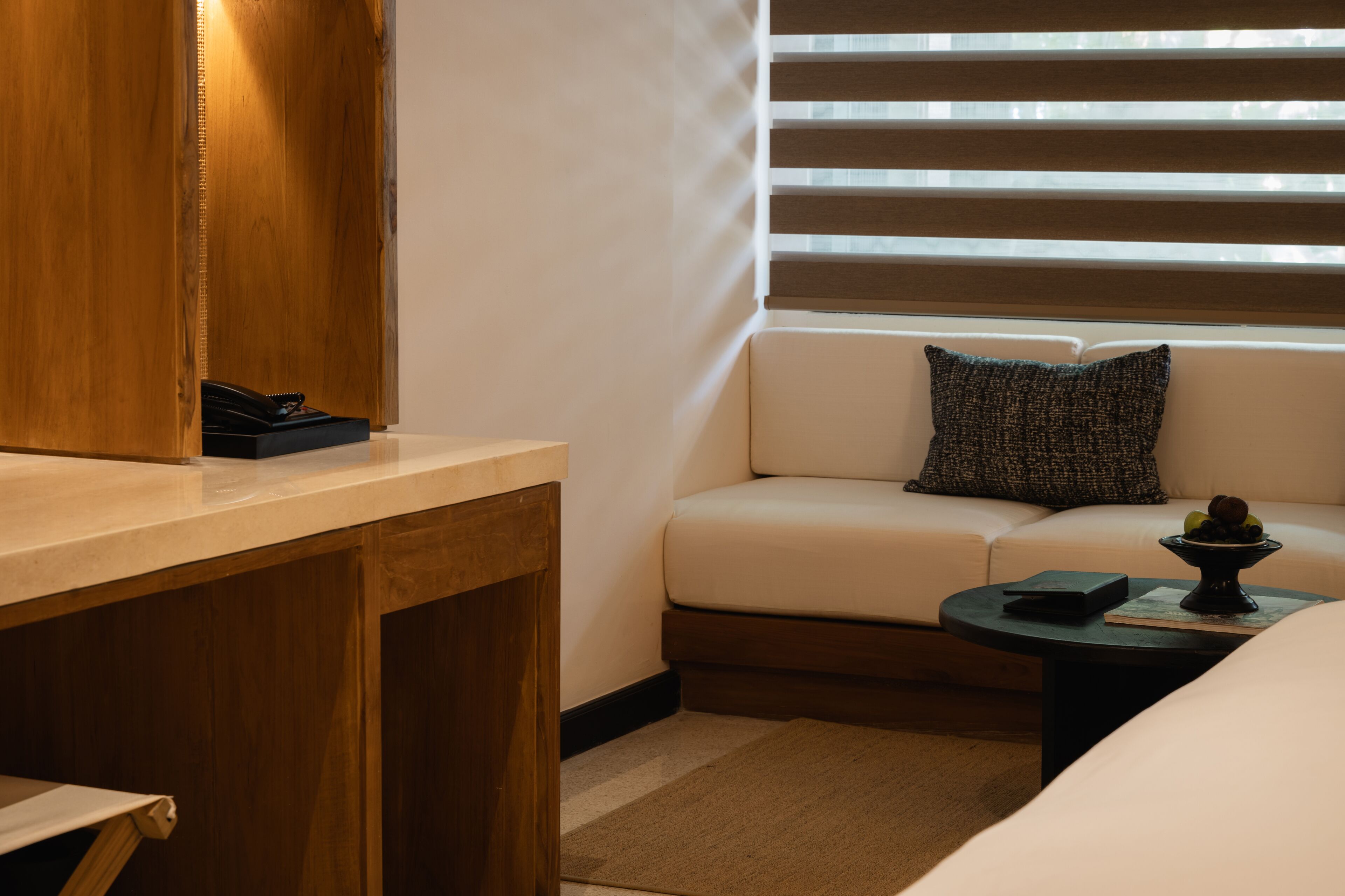 A modern room with a wooden desk, a white sofa with a textured pillow, a small round table with a book and a decorative bowl, and a window with blinds.