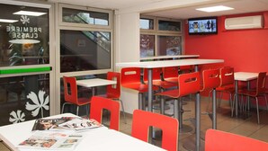 A cafeteria with red chairs and white tables, a television on the wall, and a window with a view of trees. - Première Classe Saint Malo - Saint Jouan des Guérets (Saint-Jouan-des-Guerets)