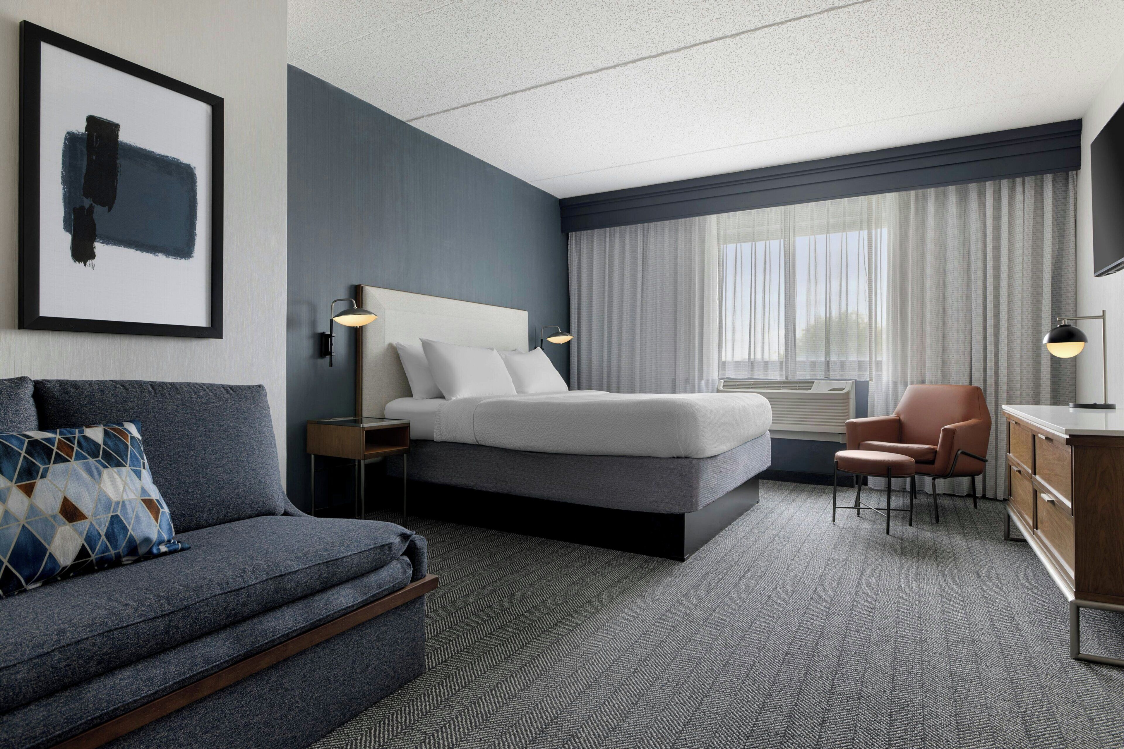 A modern hotel room with a bed, a sofa, a chair, a desk, and a large window with curtains.