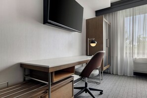 Habitación de hotel con televisor de pantalla plana montado en la pared, un escritorio con silla, armario y ventana con cortinas translúcidas.