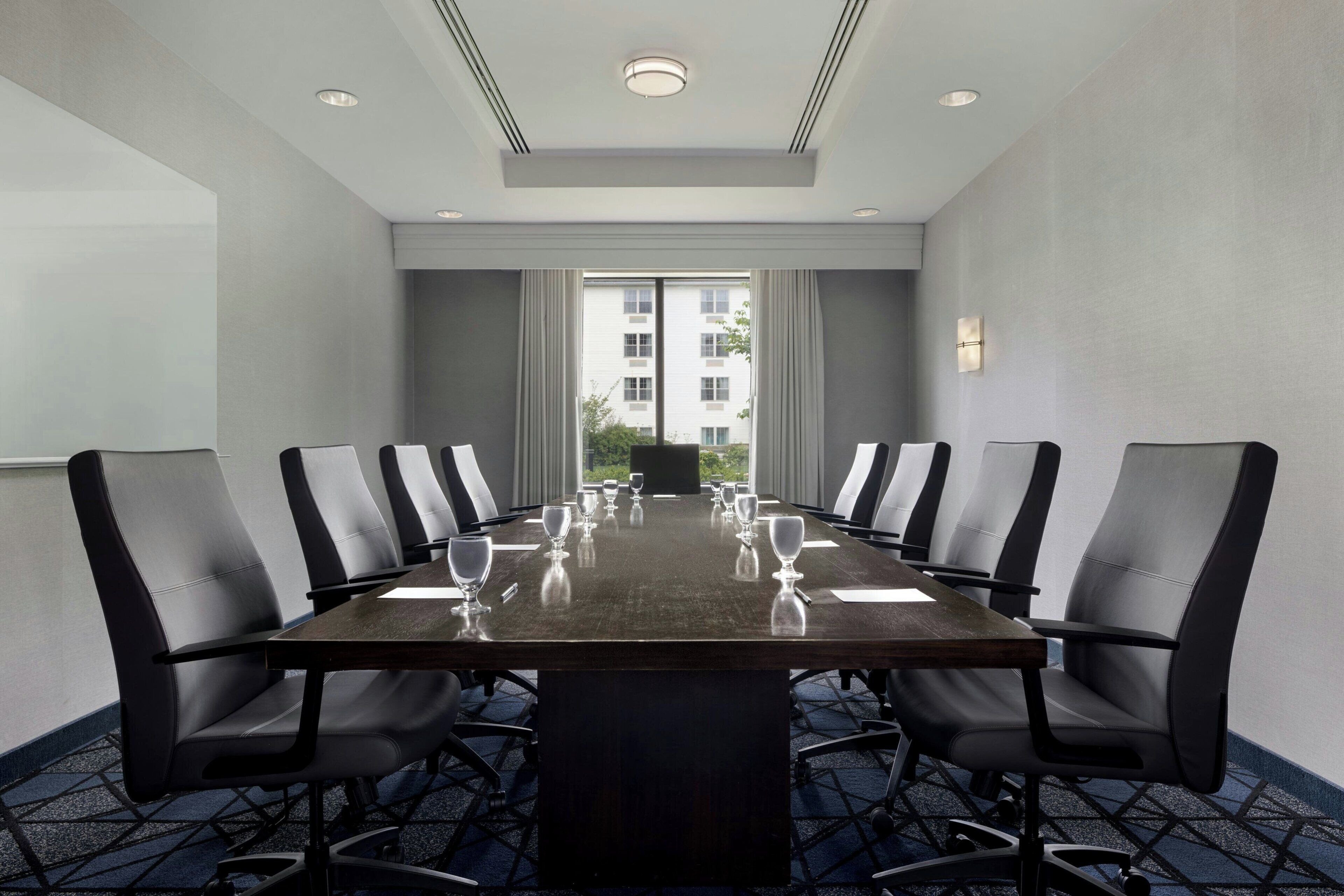 A conference room with a long table, black chairs, and a view of a building through large windows.
