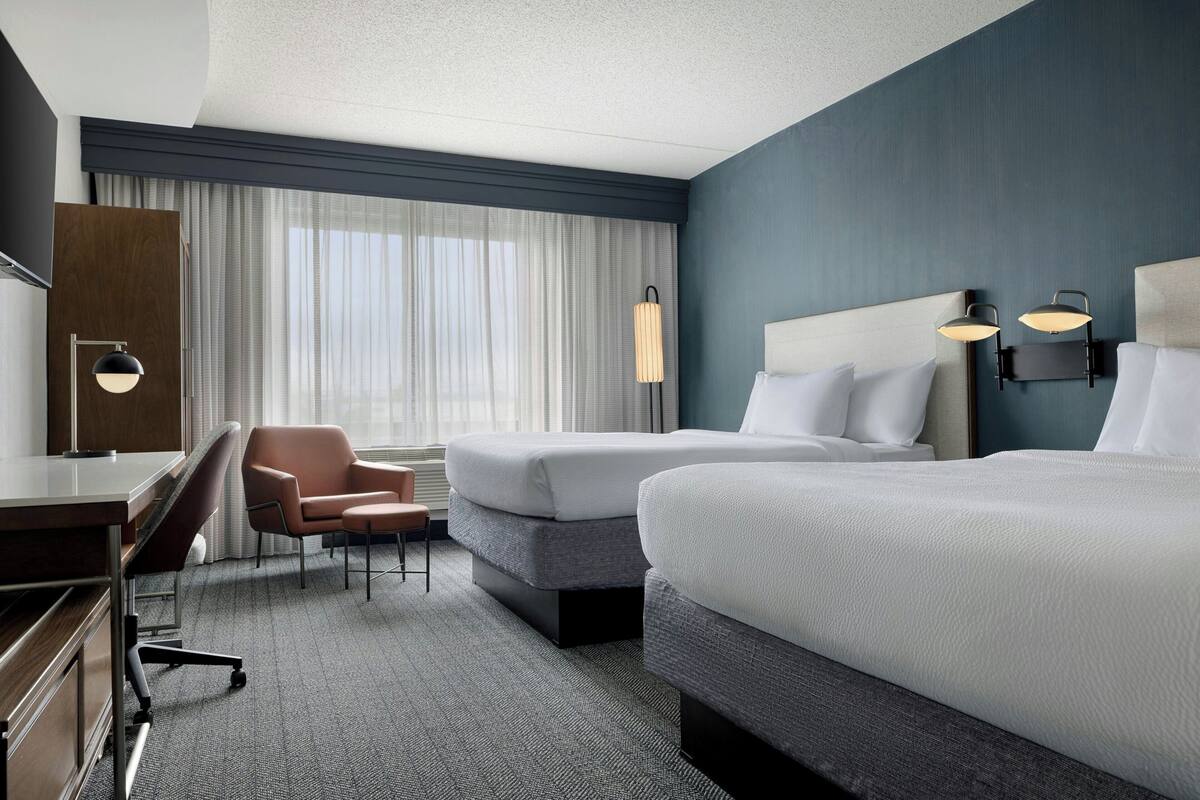 A hotel room with two beds, a desk, a chair, a lamp, and a large window with curtains.