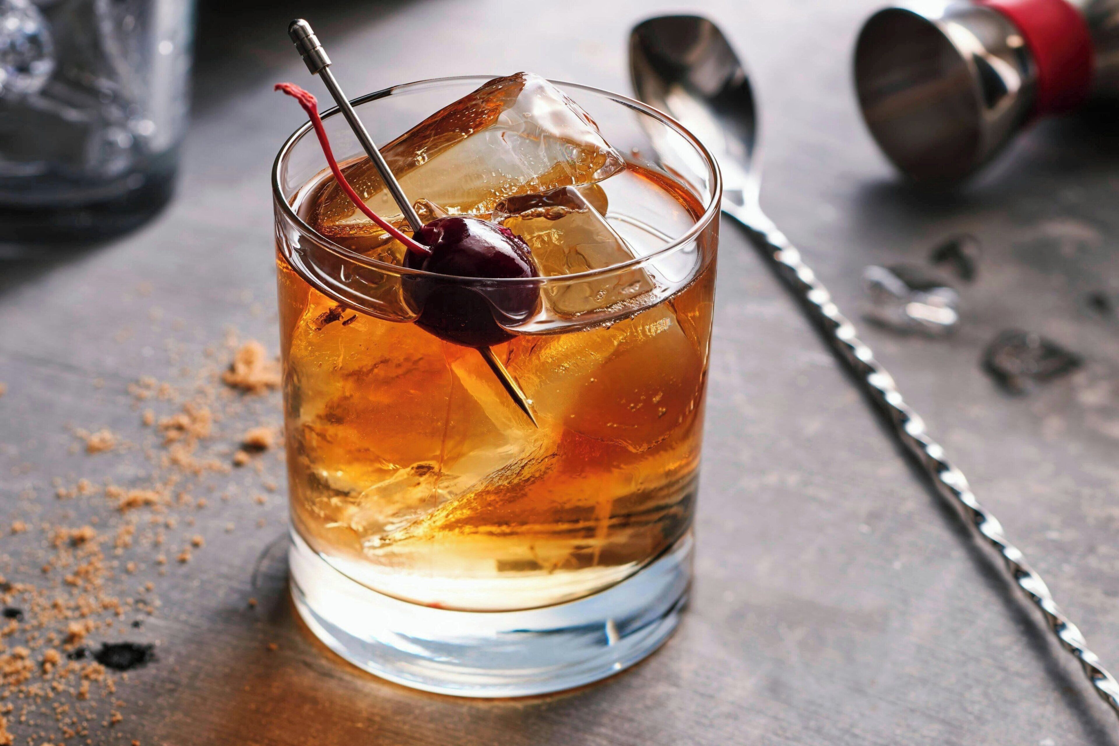 A glass with a cherry on ice, a metal straw, and a cocktail shaker in the background.